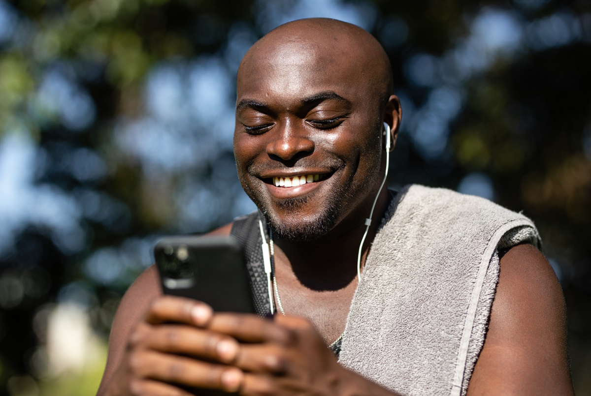 A man in exercise clothes and wearing headphones smiles while looking at a cell phone in his hands.